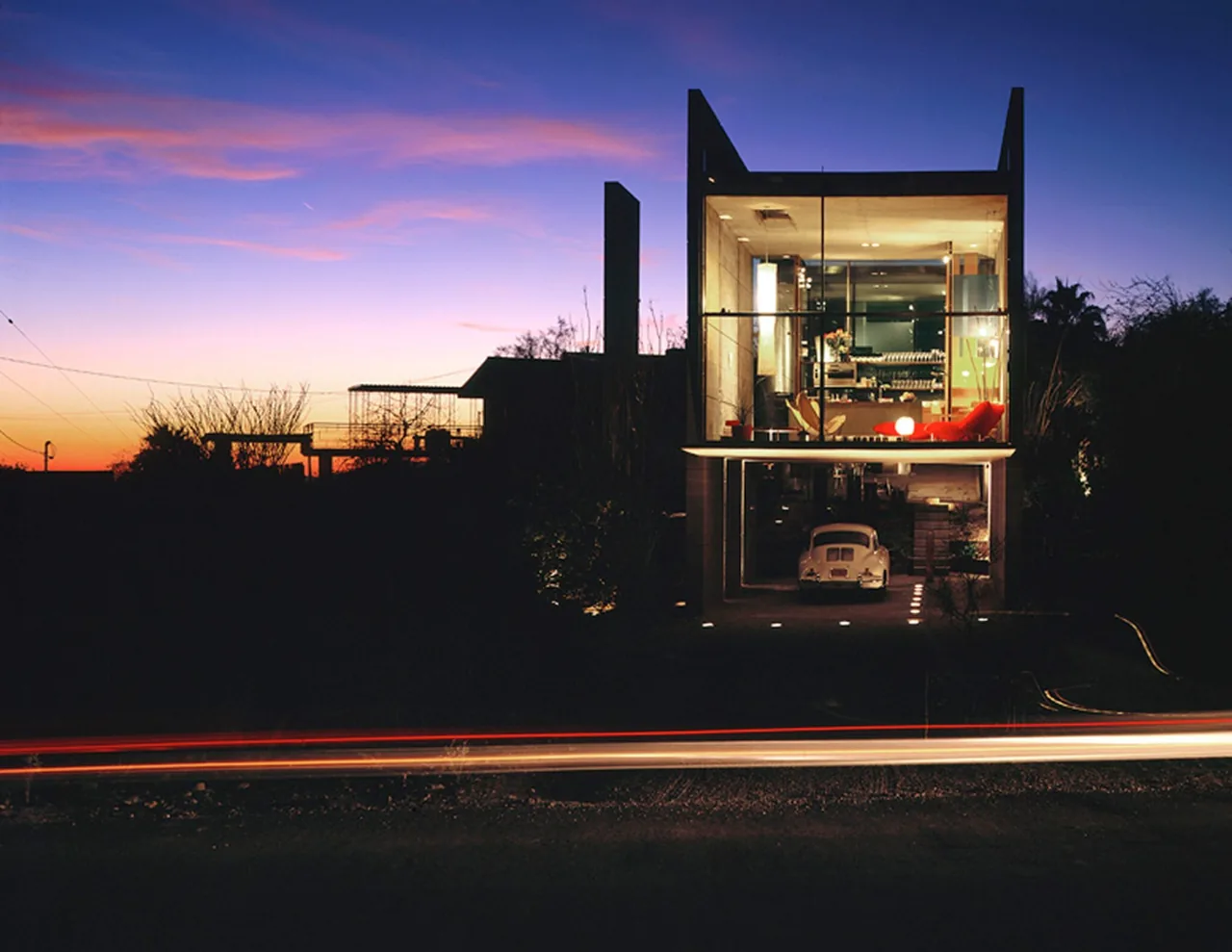 Front exterior view of a modern two-story house with floor-to-ceiling glass walls illuminated at dusk.