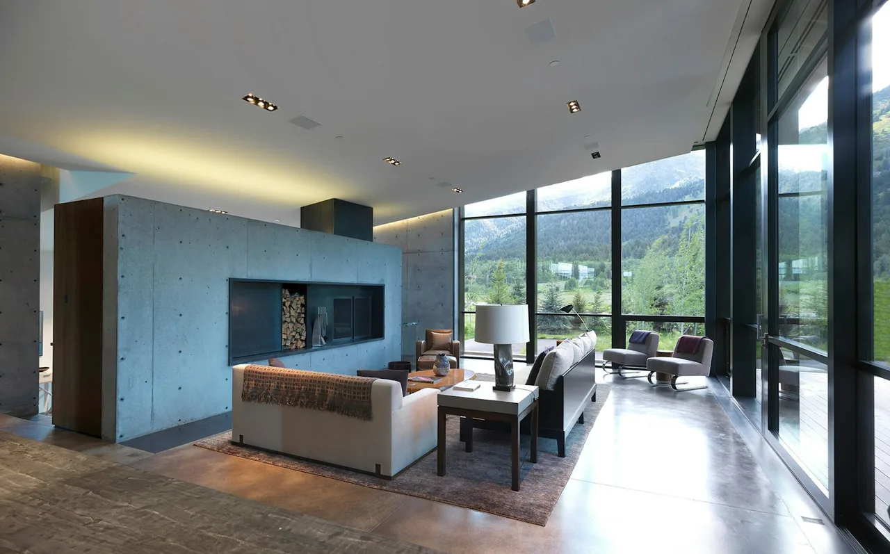 Modern living room with a central concrete fireplace and large floor-to-ceiling windows overlooking a forested mountain view.