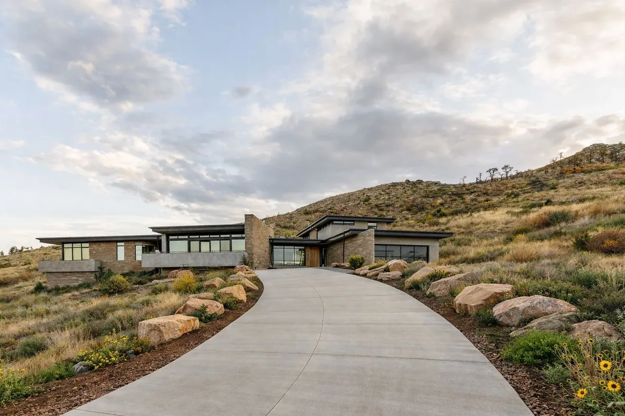 Modern single-story house nestled on a hillside with a winding concrete driveway and stone landscaping.