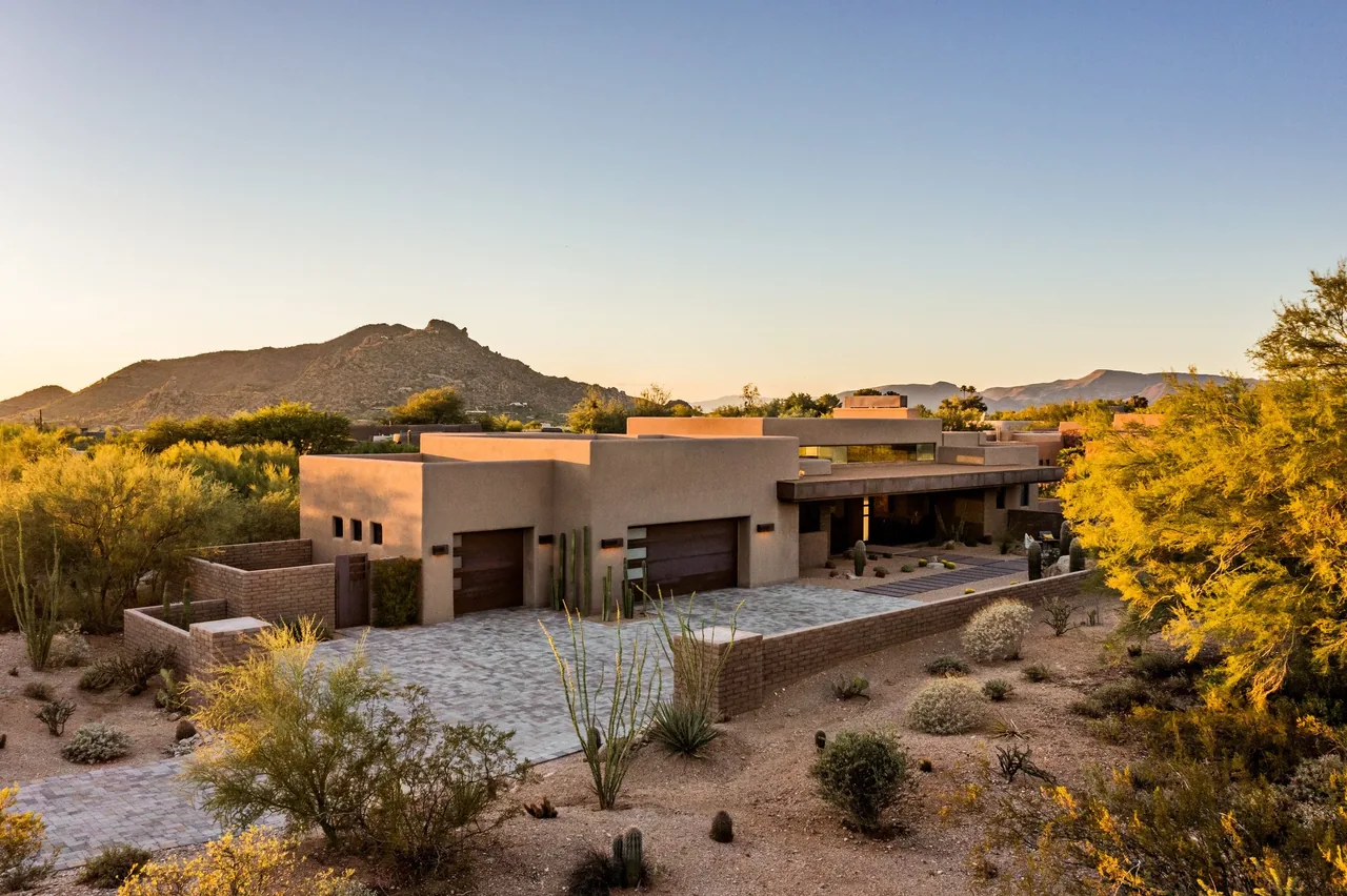 A modern flat-roof desert house with earth-tone stucco walls and a paved driveway, set against a mountain backdrop.