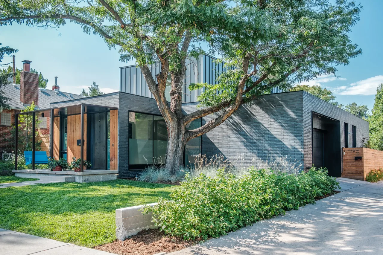 Modern single-story home with dark brick exterior, large tree in front yard, and wooden porch area with glass walls.
