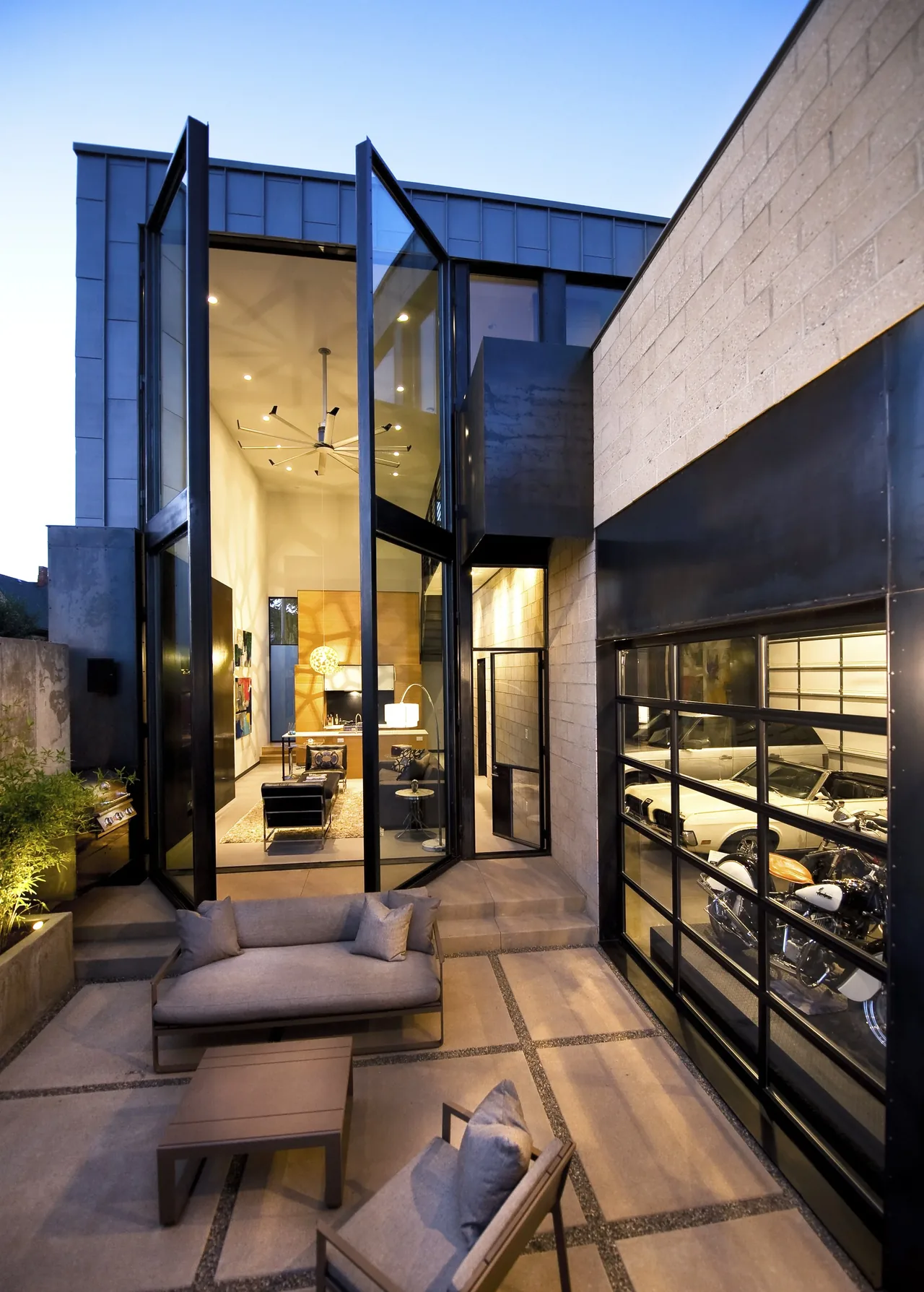 Outdoor patio seating area adjacent to a glass-walled modern home with views into a living room and a garage with a motorcycle and car.