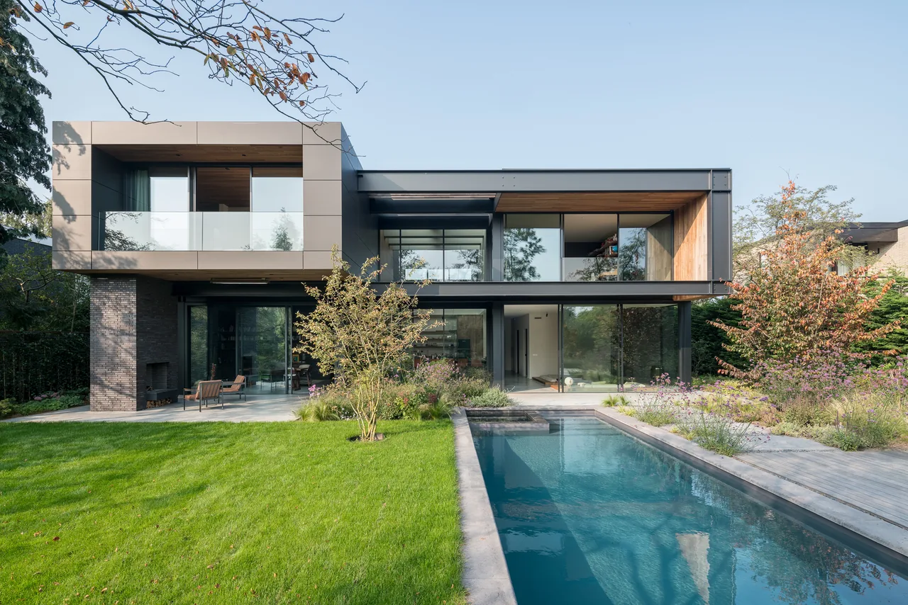 Modern two-story house with large glass windows, a patio seating area, and a swimming pool in the garden.