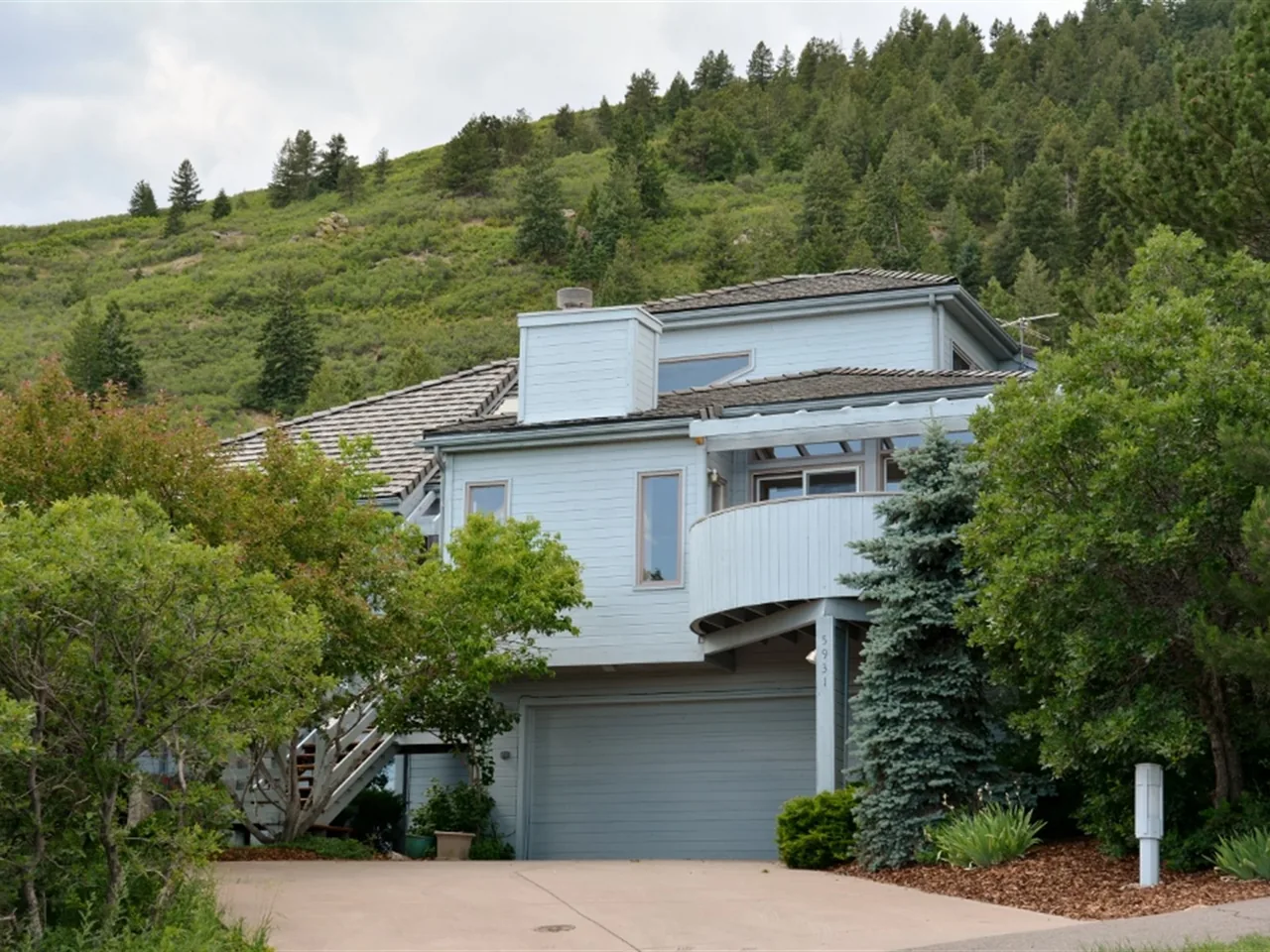Light blue multi-story house with a double garage and curved balcony surrounded by green trees on a sloping hill.
