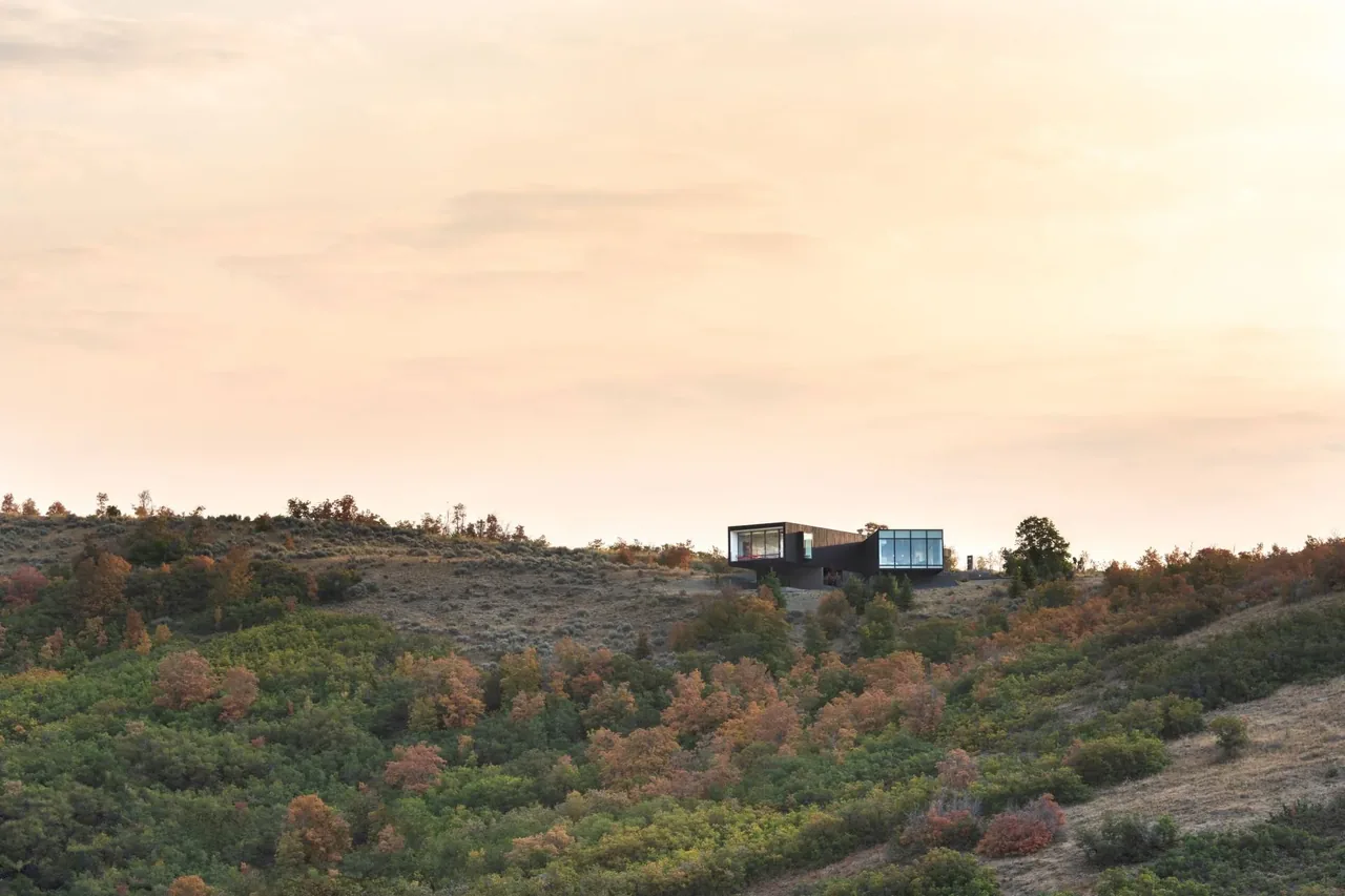 Modern house with large windows situated atop a hilly landscape with mixed green and red foliage under a cloudy sky.