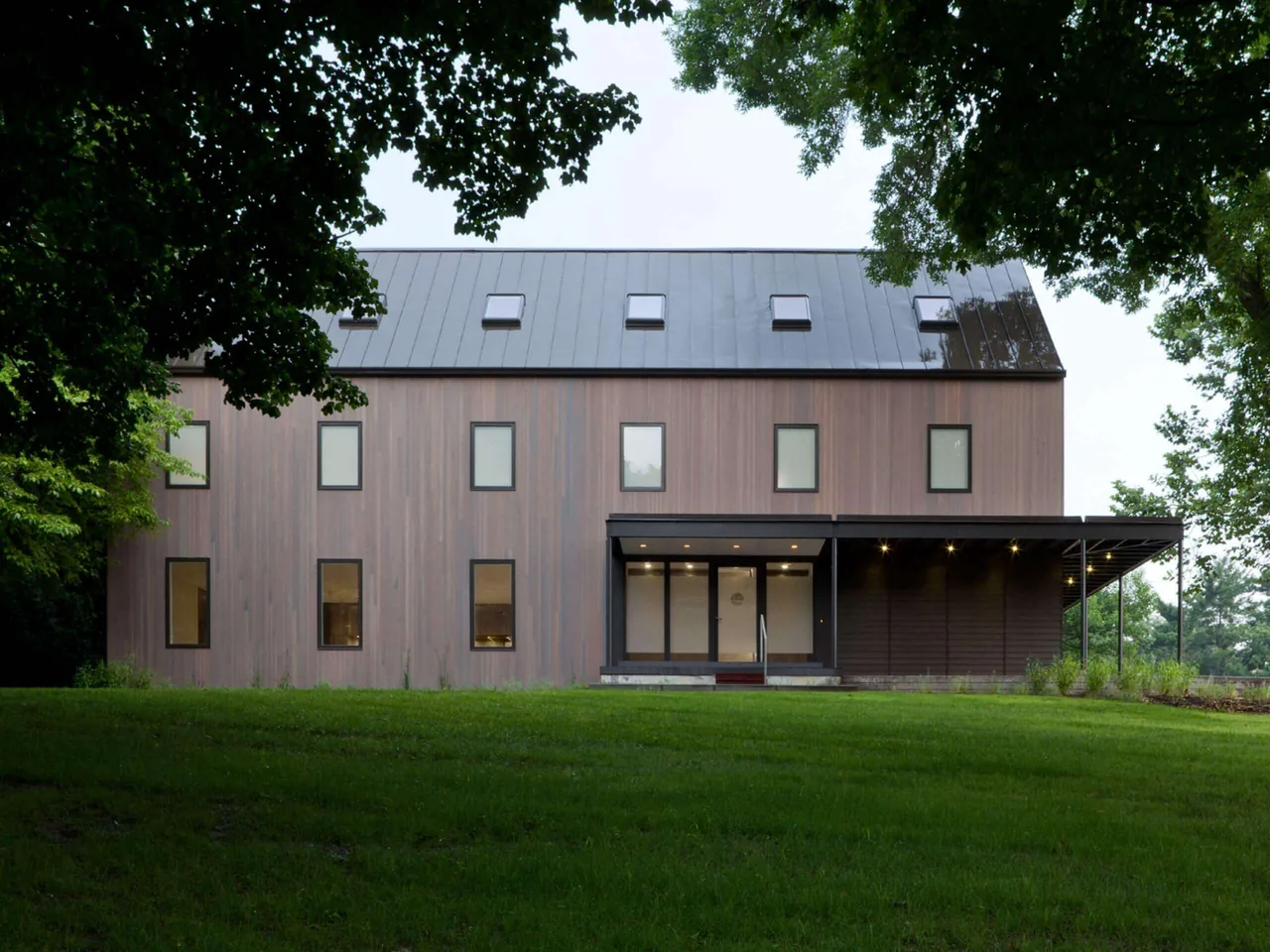 Front view of a modern two-story house with vertical wood siding and a metal gable roof, surrounded by trees and grass.