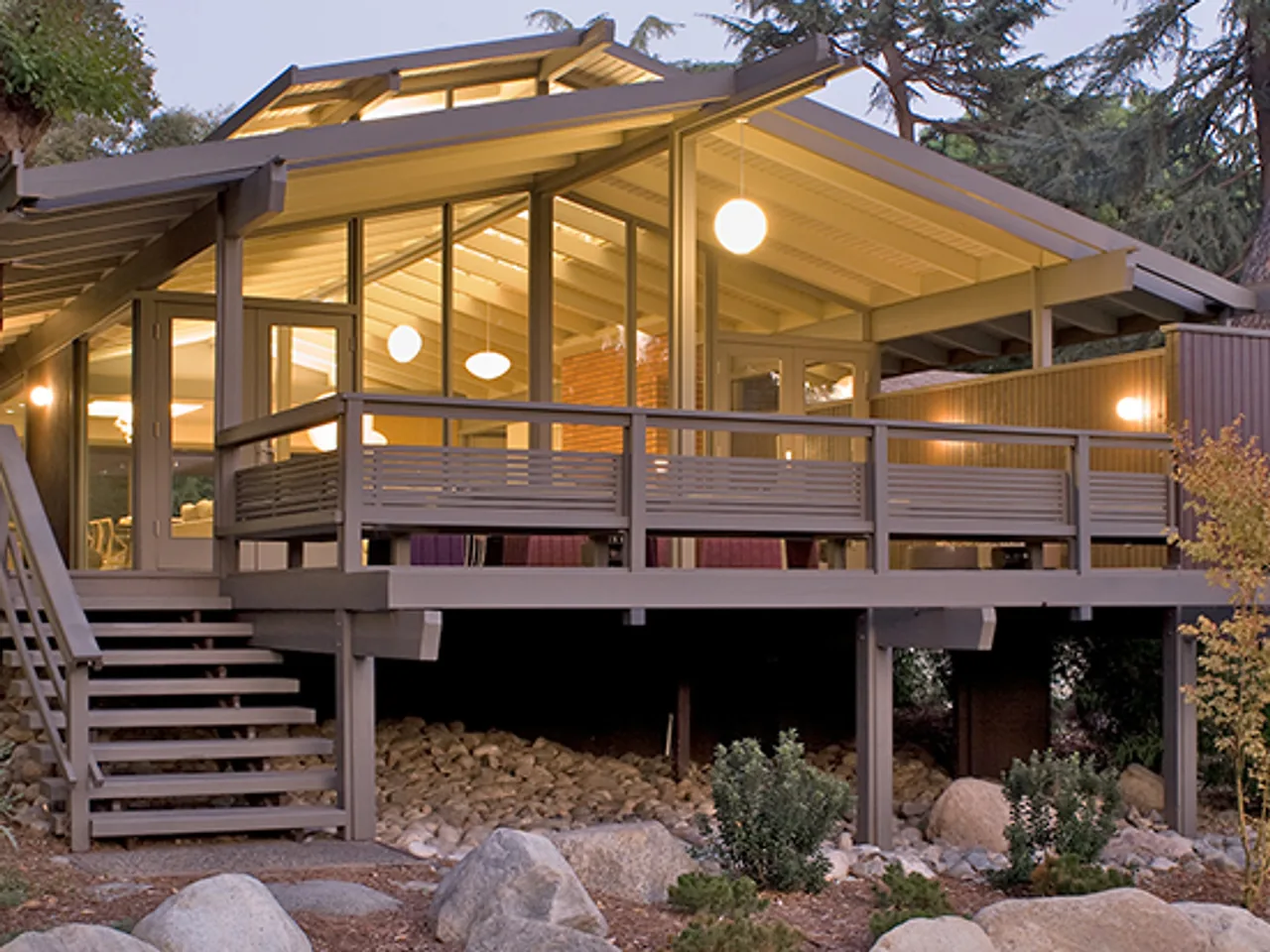 Exterior view of a midcentury modern home with large floor-to-ceiling glass windows and a covered deck raised on stilts.