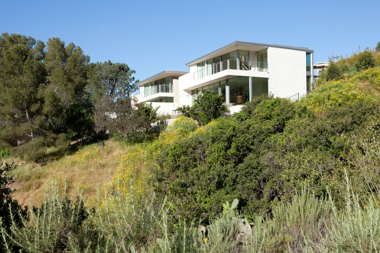 Modern white residences with large glass windows situated on a hillside with natural vegetation and blue sky.