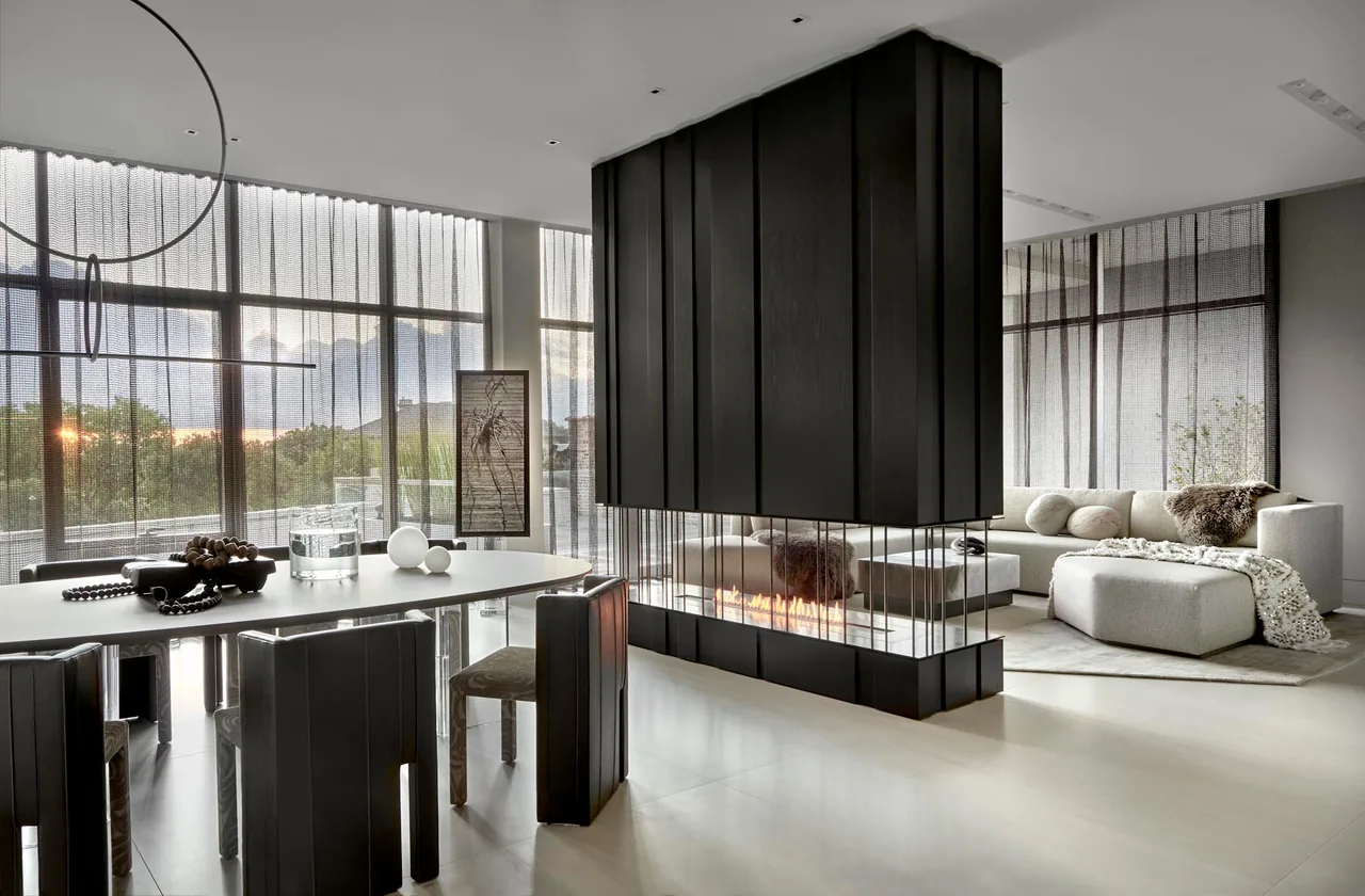 Modern open living and dining area with floor-to-ceiling windows, a suspended black fireplace, and neutral furnishings.