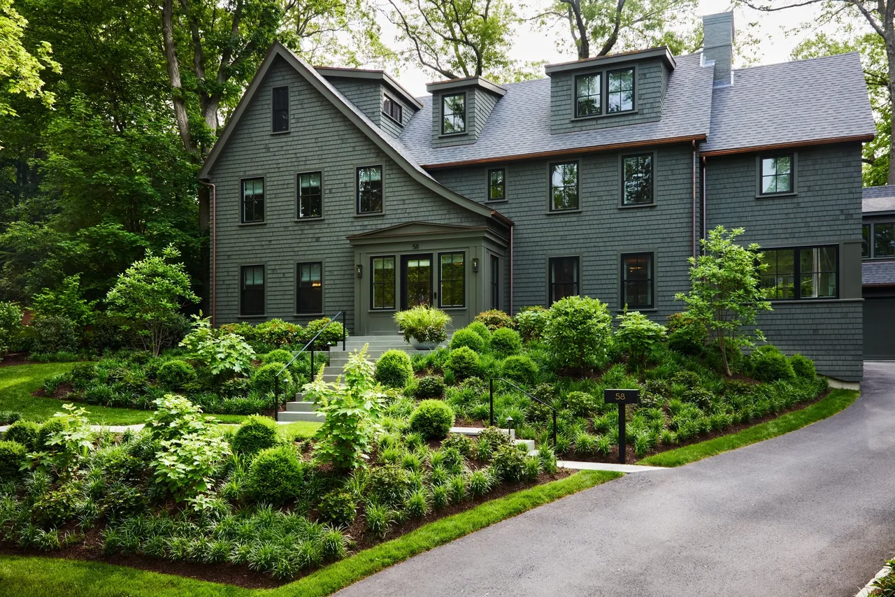 Gray shingle exterior house with multiple dormer windows, surrounded by lush greenery and a curved driveway.