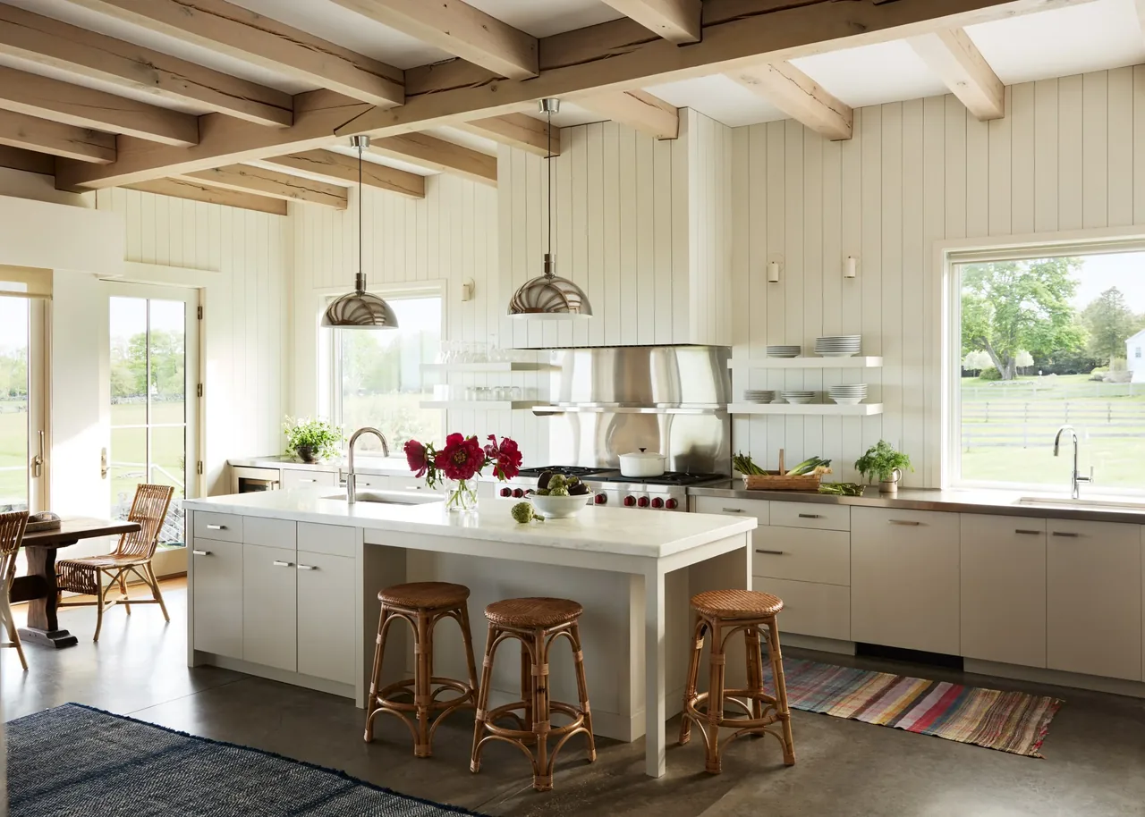 Bright kitchen with white cabinetry, wooden beams on ceiling, and three wicker stools at a large island under metal pendant lights.