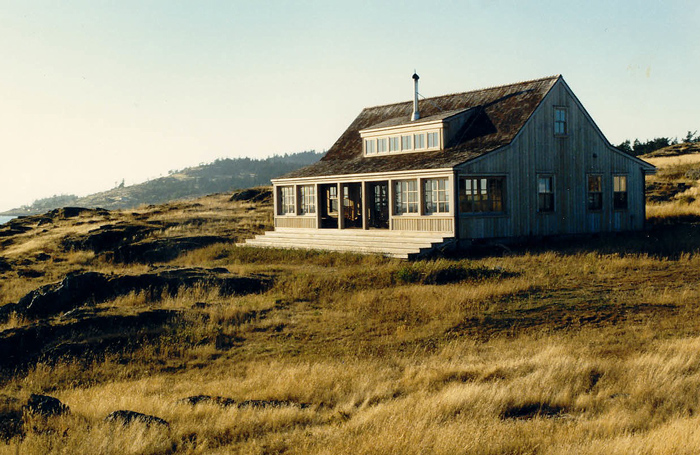 A wood-sided house with multiple windows and a covered front porch sits on a grassy hillside near the coast.