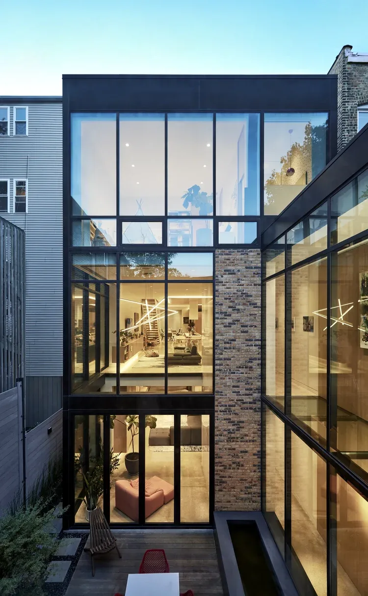 View of a modern three-story house facade with large floor-to-ceiling black-framed windows and a brick wall section, overlooking a small, w(