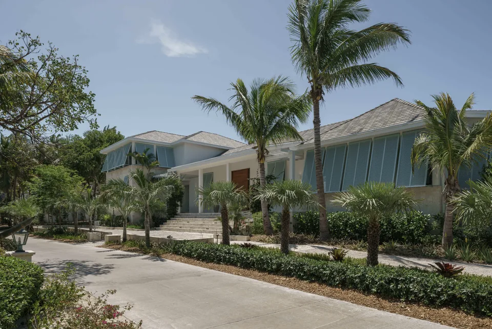 Front view of a modern coastal house with palm trees and a wide concrete driveway under clear sky.