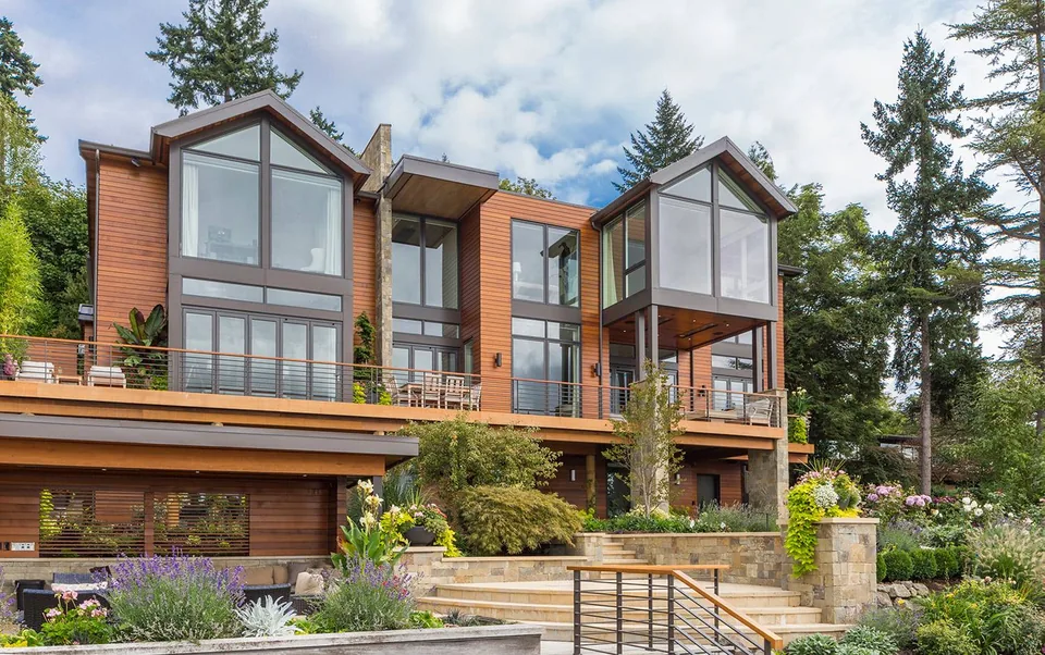 Exterior view of a modern multi-level house with large glass windows and wood siding, surrounded by a landscaped garden.