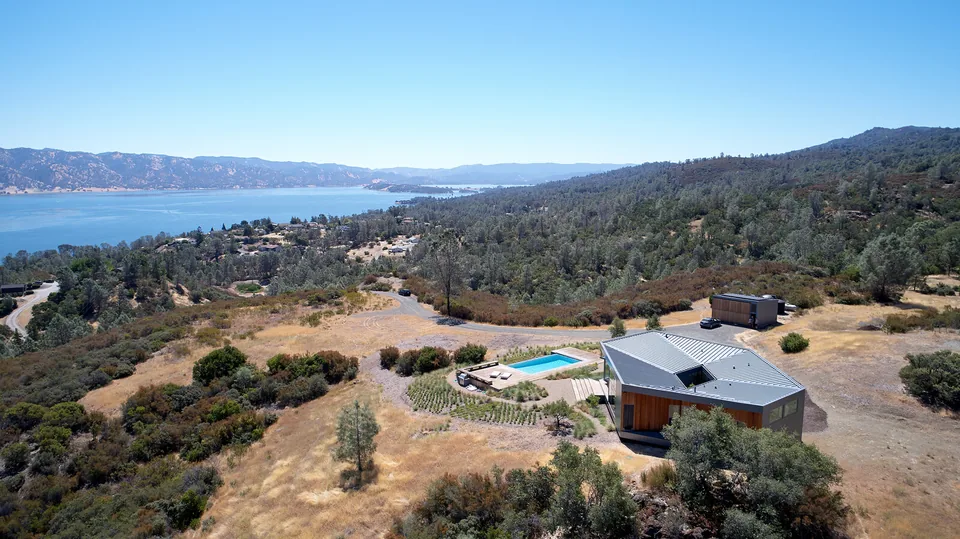 A modern house with a metal roof and wooden siding on a hillside overlooking a large body of water and forested mountains.
