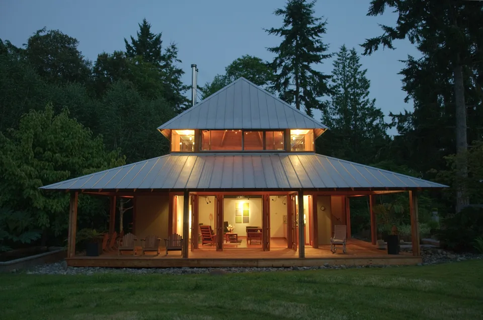 Evening view of a metal-roofed house surrounded by trees with a wrapped porch and warm interior lights.