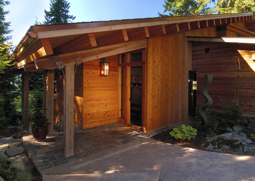 Covered entryway of a house with warm wood siding and slate flooring in a wooded setting.